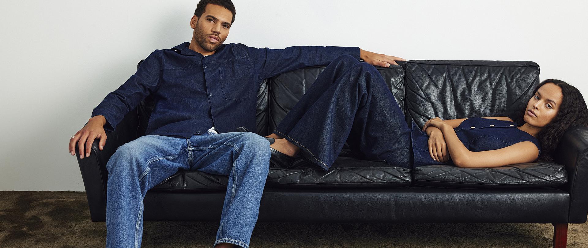 A man and a woman are lounging on a sofa, wearing denim outfits.